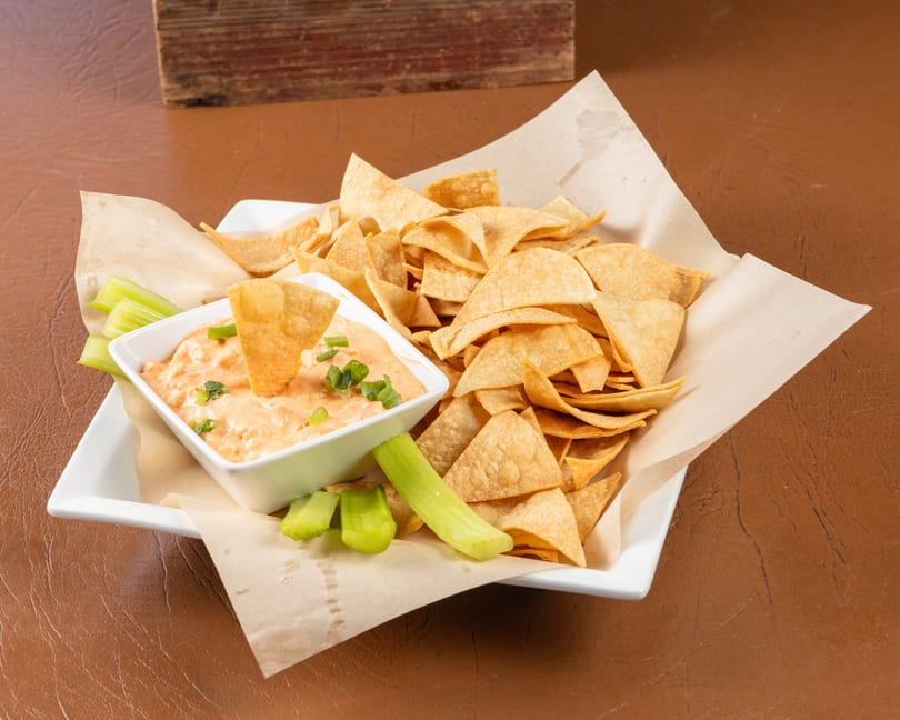 A white platter holds a bowl of creamy dip with green onions, tortilla chips, and celery sticks.