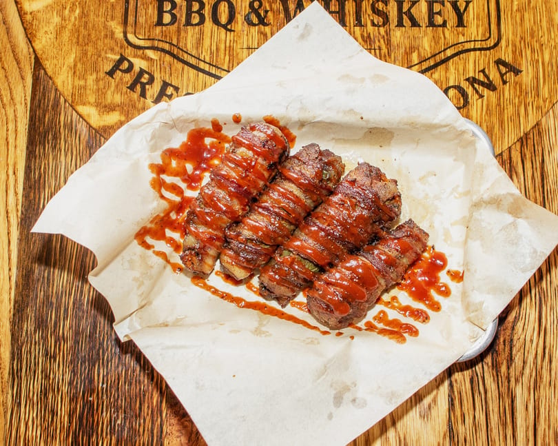 Four bacon-wrapped jalapeño poppers drizzled with BBQ sauce on parchment paper, on a wooden barrel.