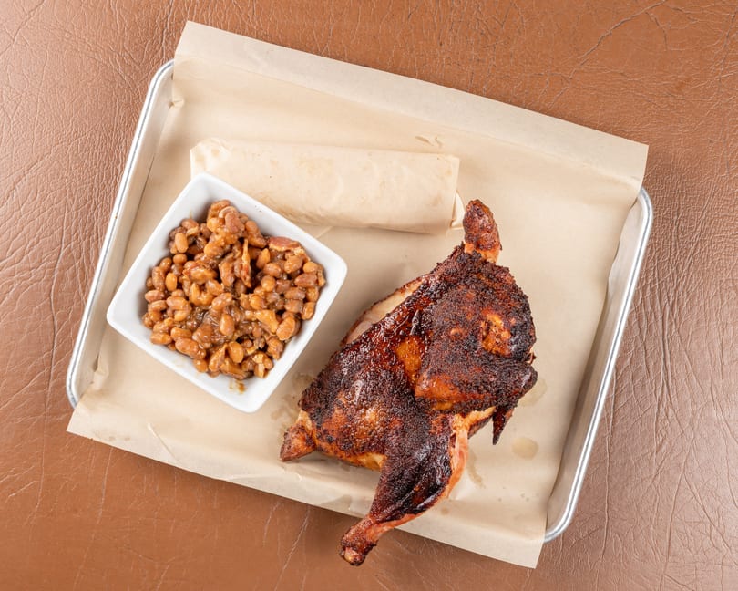Half a roasted chicken with crispy skin and a side of baked beans on a metal tray.