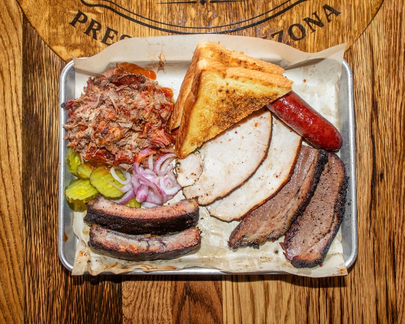 A metal tray with pulled pork, ribs, brisket, turkey, sausage, pickles, onions, and toast.