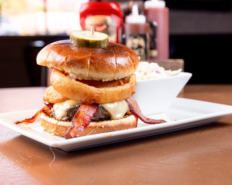 A gourmet burger with bacon, cheese, onion rings, and a pickle, served with coleslaw.