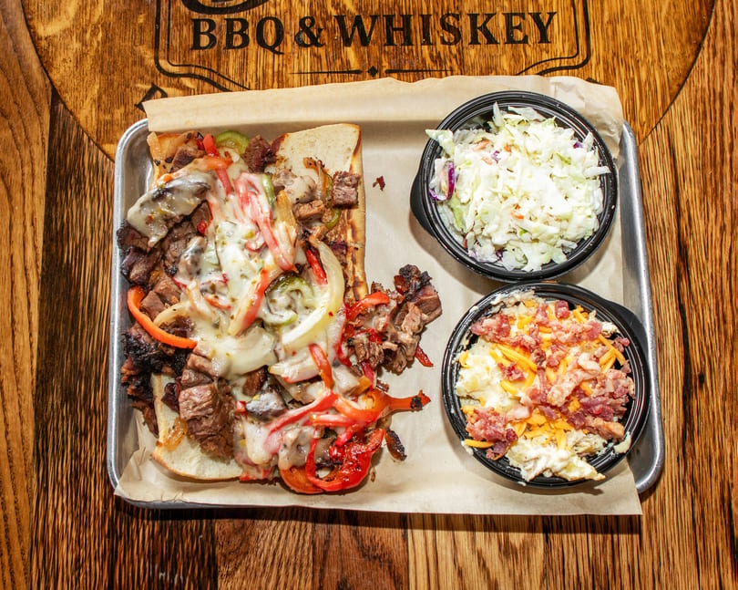 Cheesesteak sandwich with peppers and onions, coleslaw, and loaded mashed potatoes on a tray.