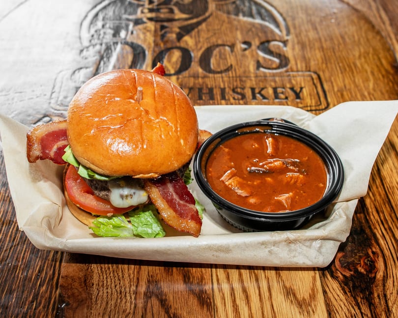 Bacon cheeseburger with a side of chili on a wooden table with "Doc's Whiskey" carved into it.