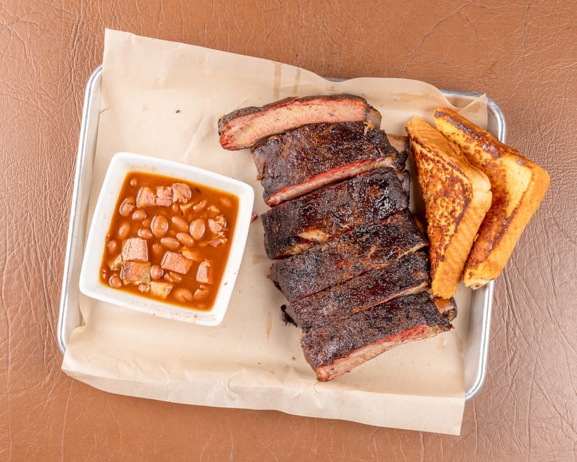 A tray of BBQ ribs, baked beans, and two slices of Texas toast on a brown background.