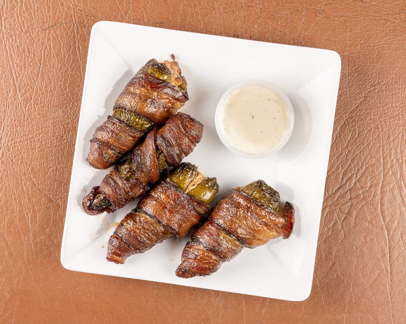 Four bacon-wrapped jalapeño poppers with a side of ranch dressing on a white plate.