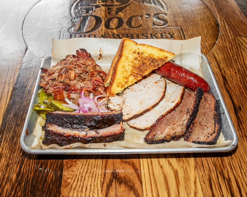 A metal tray of Doc's BBQ and Whiskey's smoked meats, including brisket, pulled pork, sausage, and turkey.
