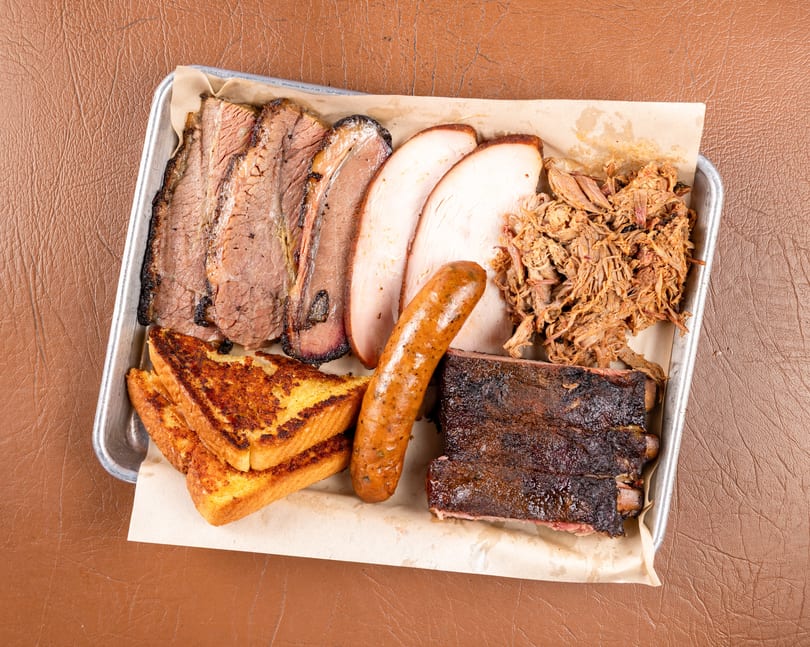 A metal tray filled with various barbecued meats, including brisket, turkey, pulled pork, sausage, ribs, and toast.