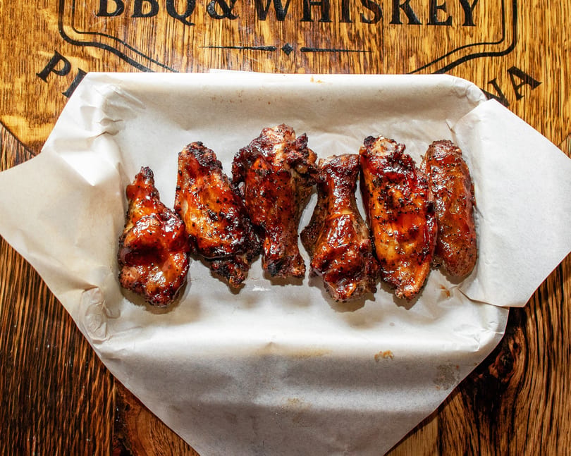 Six glazed BBQ chicken wings in a paper-lined basket on a wooden table with a BBQ logo.